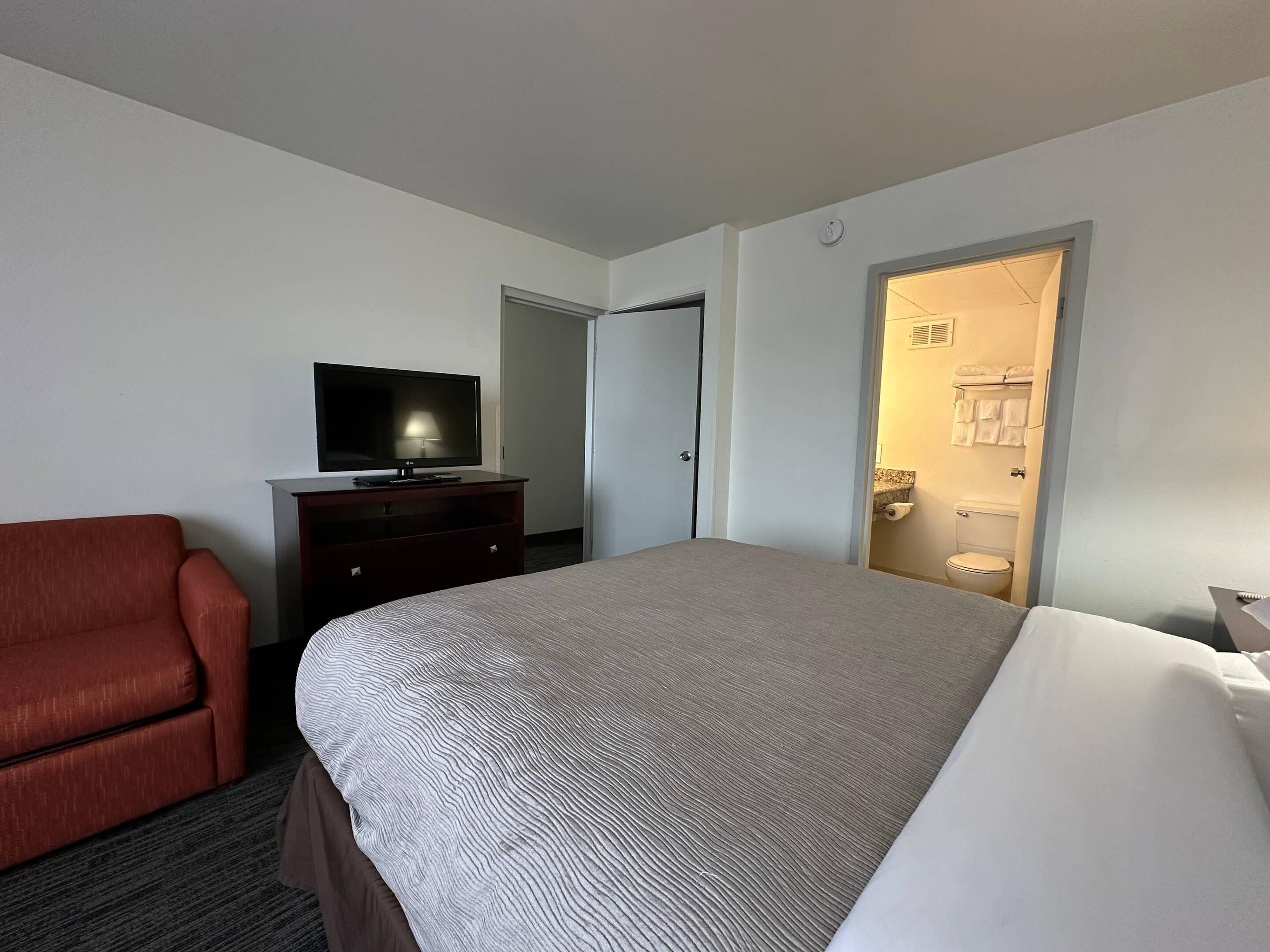 Ein Hotelzimmer mit einem Bett, einem Fernseher auf einer Kommode, einem roten Sessel und einem durchgegangene Tür erkennbaren Badezimmer.