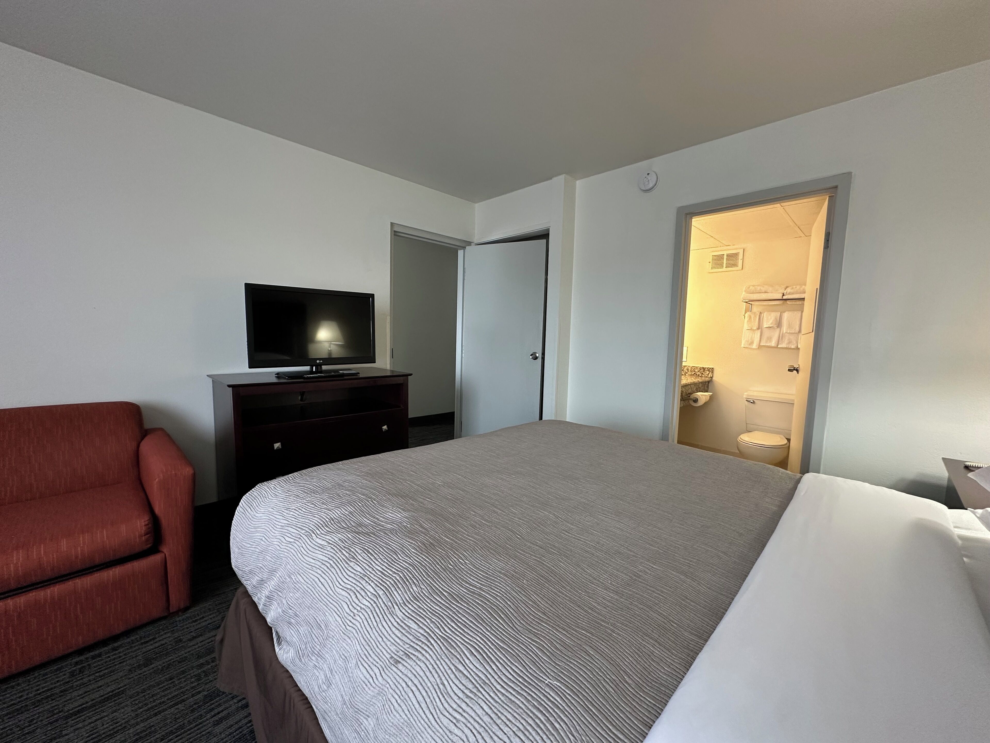 A hotel room with a bed, a TV on a dresser, a red chair, and a bathroom visible through an open door.