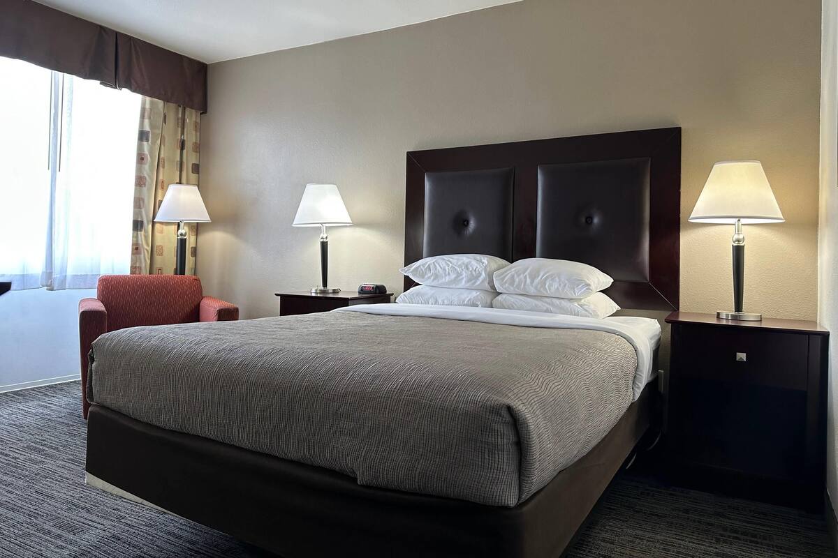 A hotel room with a large bed, two bedside lamps, a red chair, and a window with curtains.