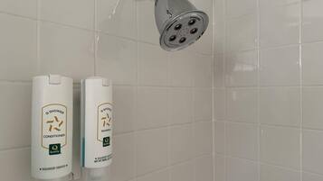 A bathroom with white tiles, a shower with multiple nozzles, and two bottles of Q Shower conditioner and shampoo.