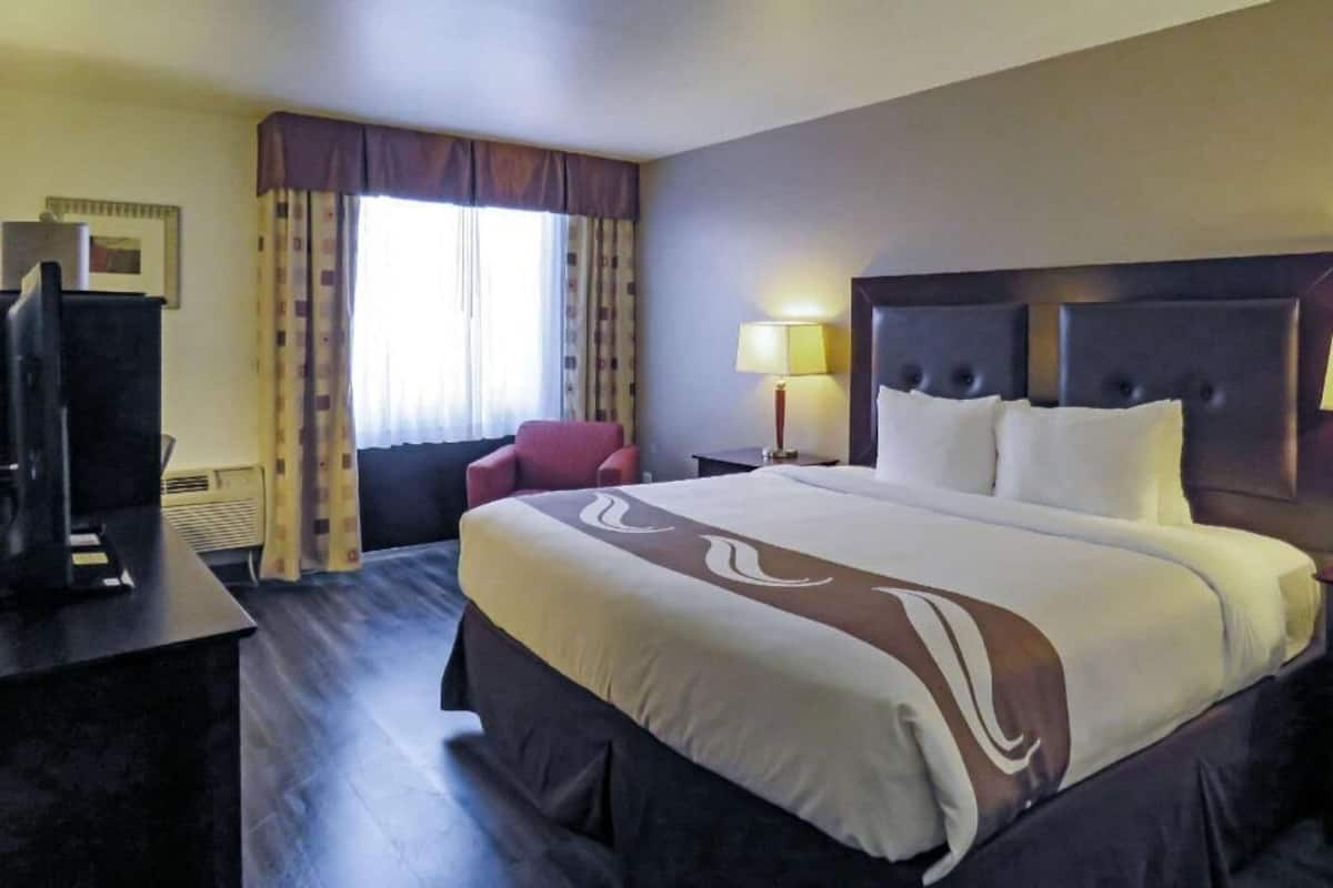 A hotel room with a large bed, a desk with a computer, a chair, a window with curtains, and a lamp.