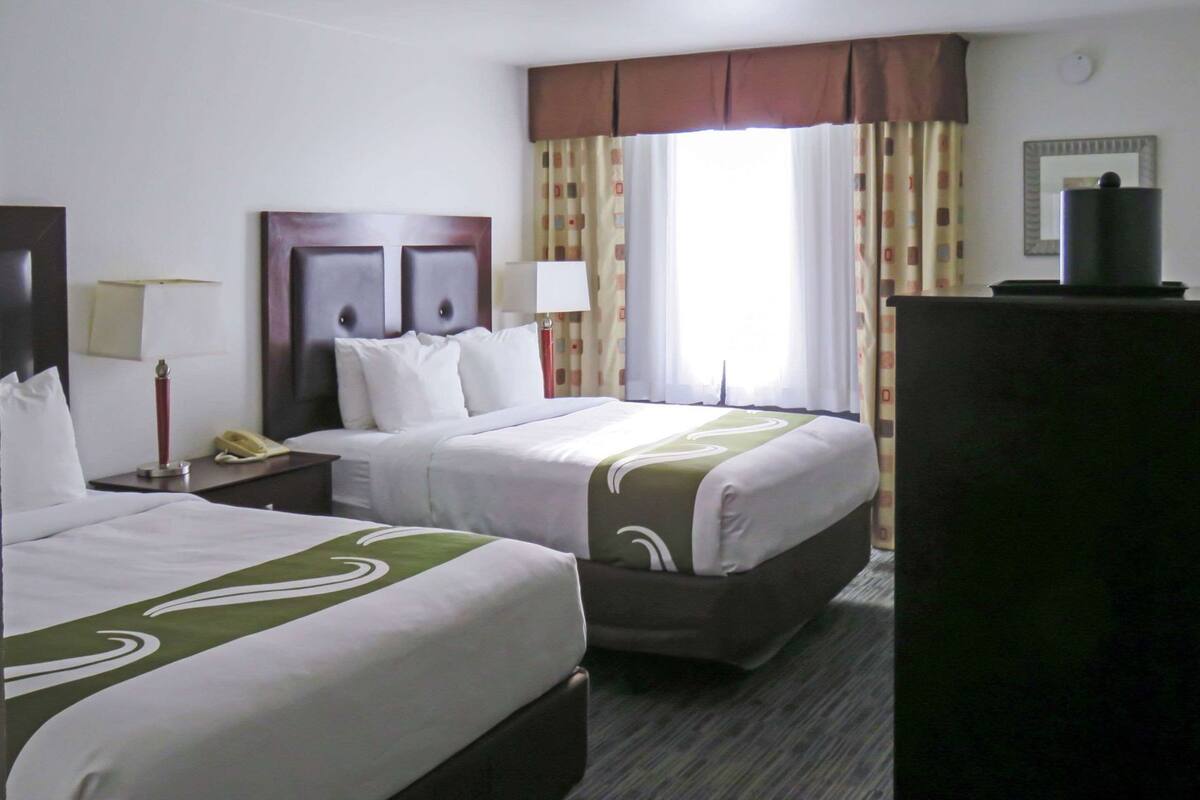 A hotel room with two beds, a nightstand with a lamp, a dresser, and a window with curtains.