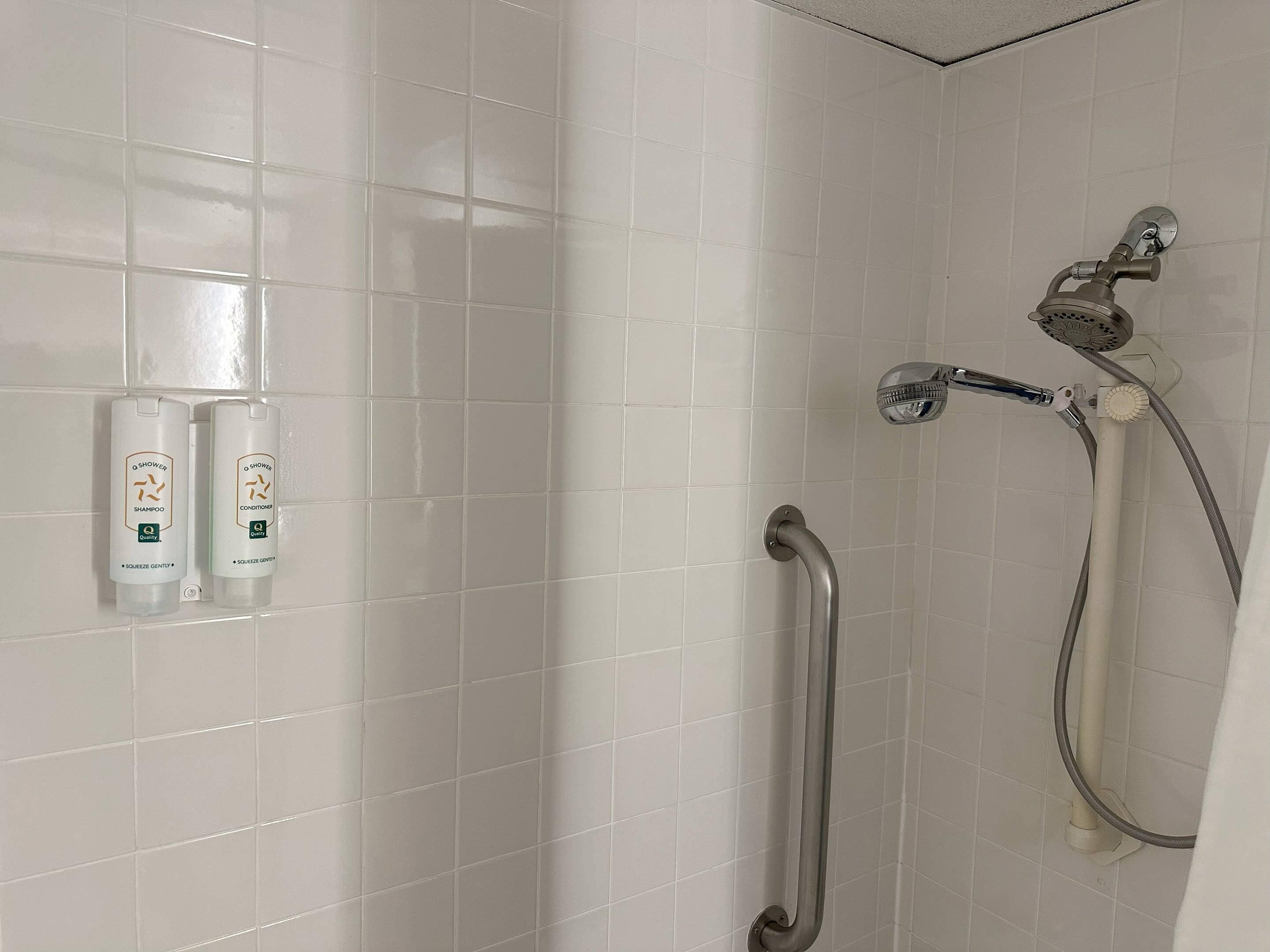 Baño con paredes de azulejos blancos, ducha con cabezal de ducha manual y dos botellas de champú y acondicionador O Shower.