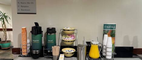 Mostrador de desayuno con dispensadores de café, jarras de jugo y un cartel que indica la ubicación de habitaciones y recepción.