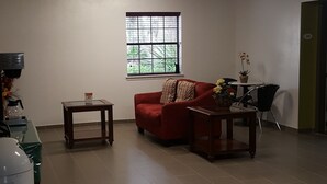 A spacious room with a red sofa, a small table, a window with blinds, and a potted plant. - Suburban Studios Tallahassee near University (Tallahassee)