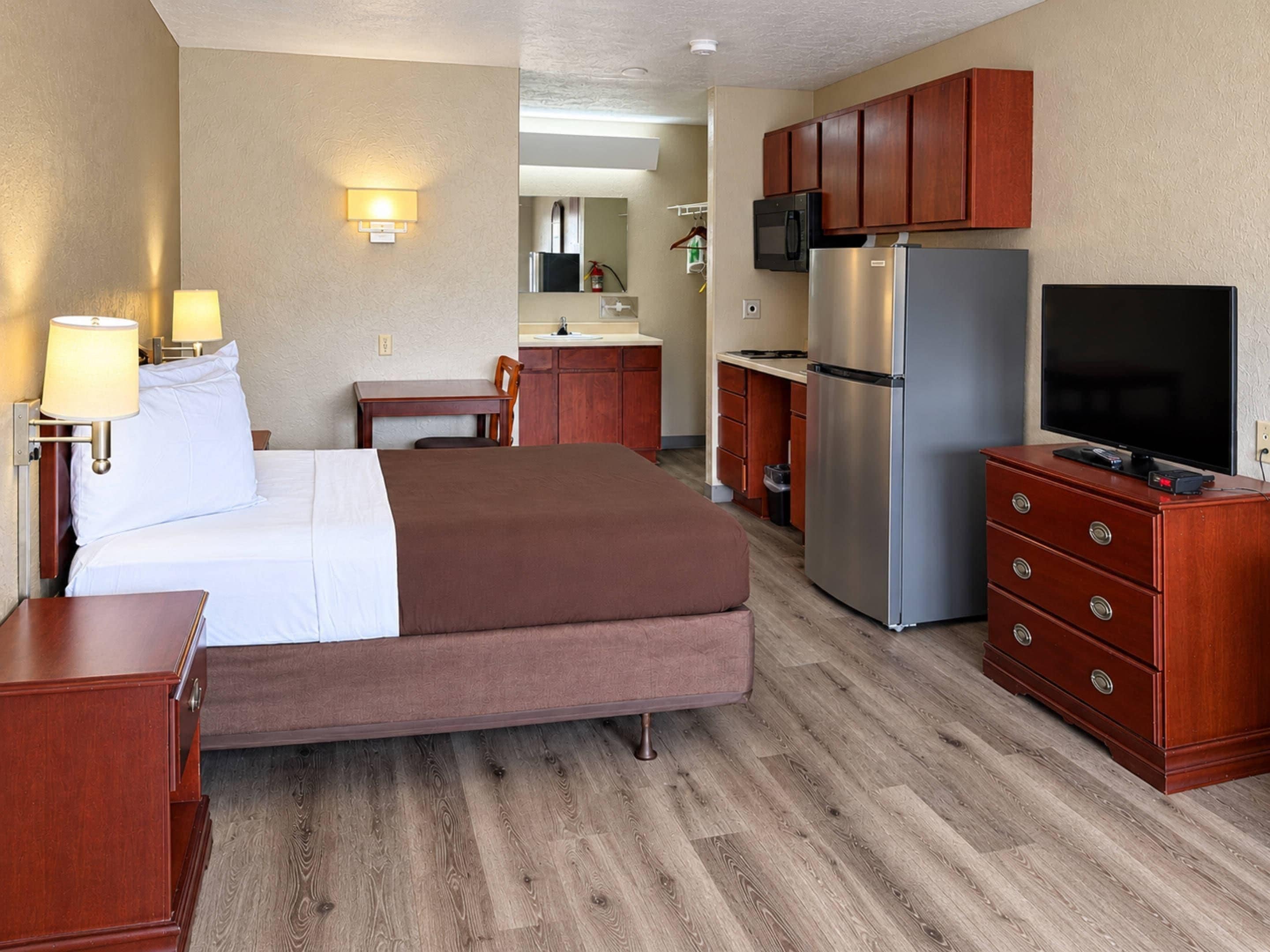 A hotel room with a bed, a desk, a kitchenette, and a television.