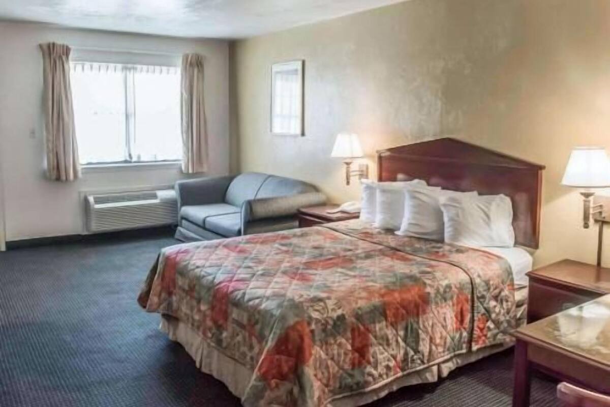 A hotel room with a bed, a chair, a desk, a lamp, and a window with curtains.