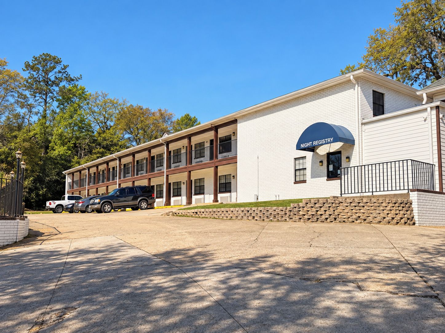 A two-story motel with a night registry sign, a parking lot, and a stone wall.