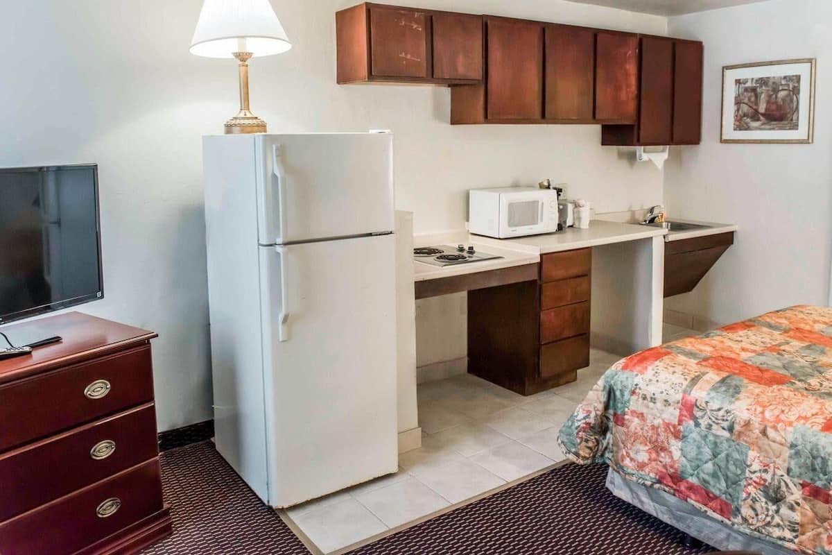 A compact hotel room with a kitchenette, a bed, a TV, and a lamp.