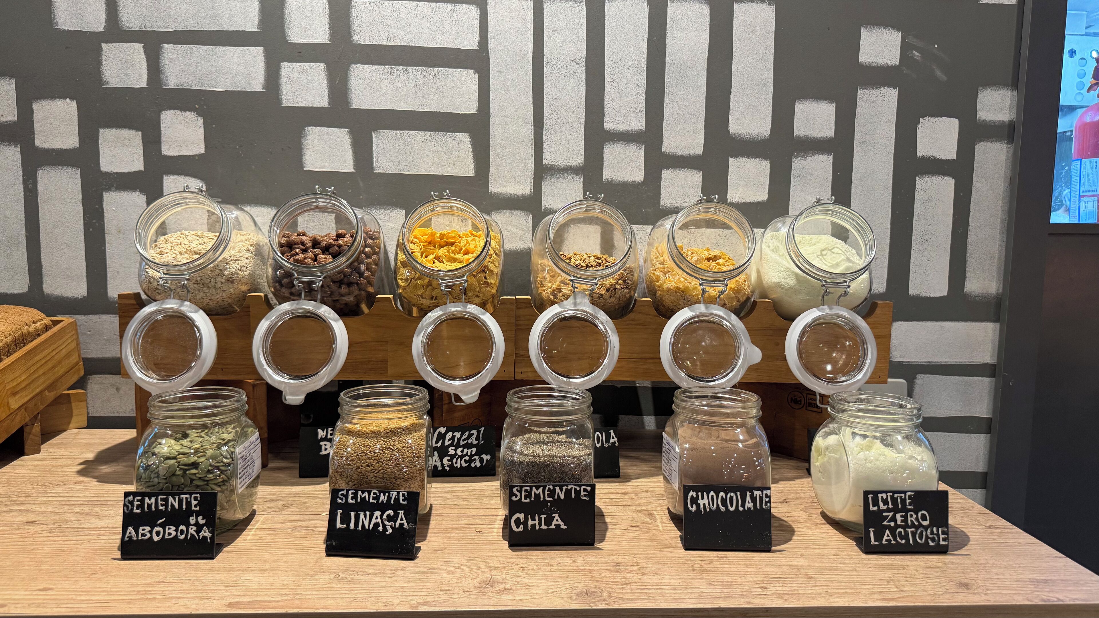 Exhibició de diferents aliments en pots de vidre amb etiquetes, disposats sobre una estant de fusta davant d'una paret amb un disseny geomètric.