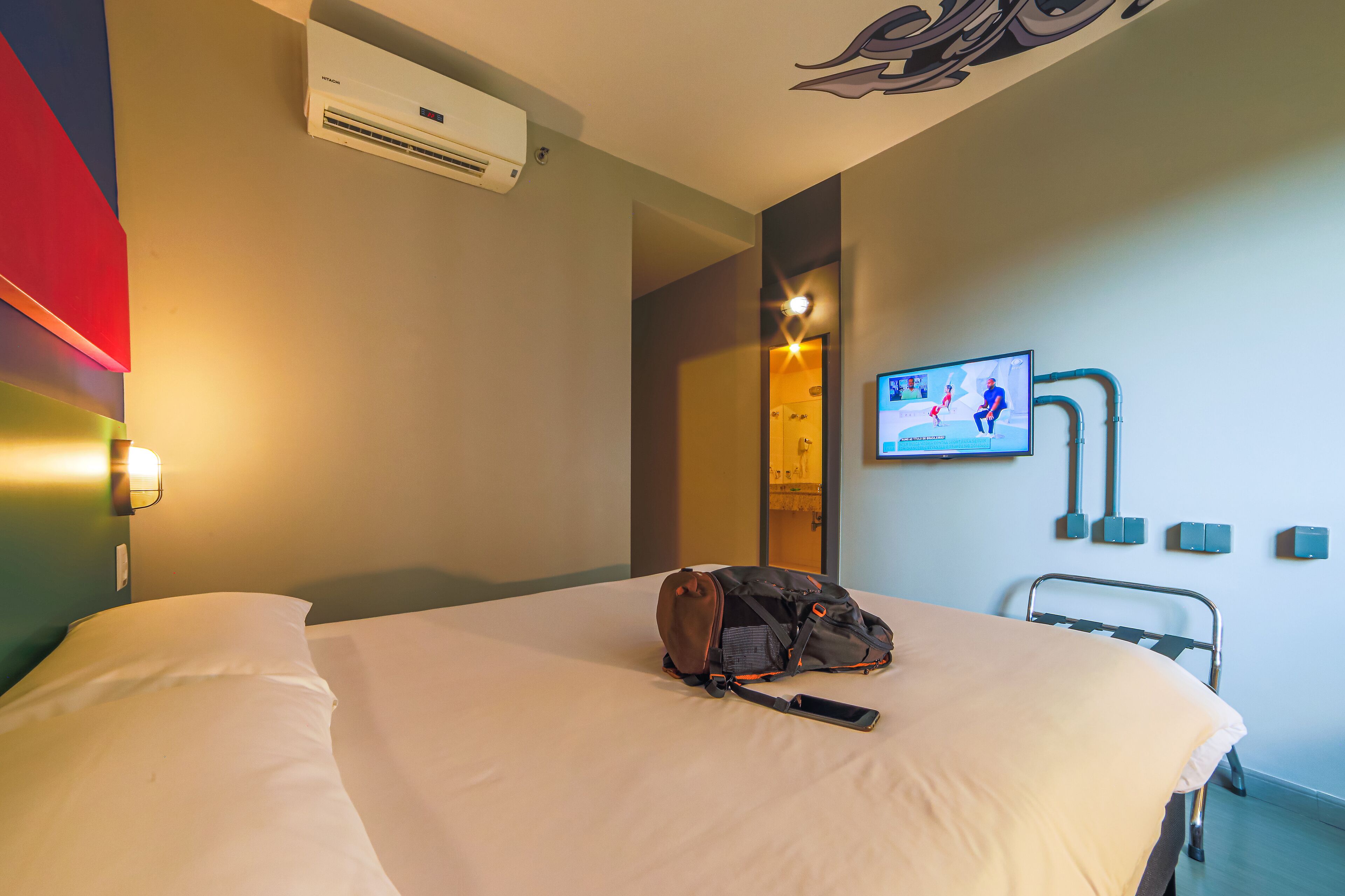 A hotel room with a bed, a wall-mounted TV, an air conditioner, and a bedside table.