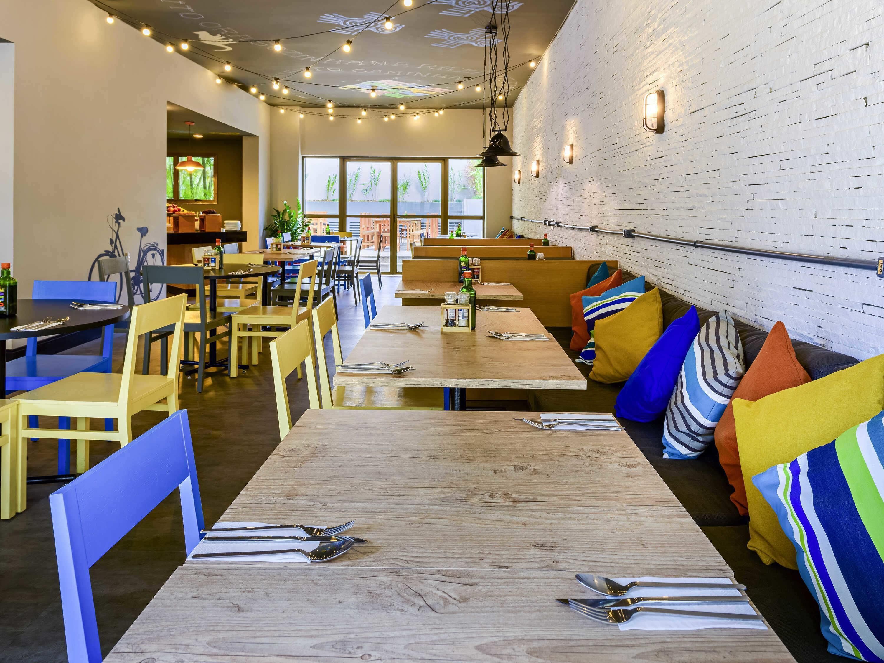 A modern restaurant with wooden tables and blue chairs, a brick wall, and a view of greenery outside.