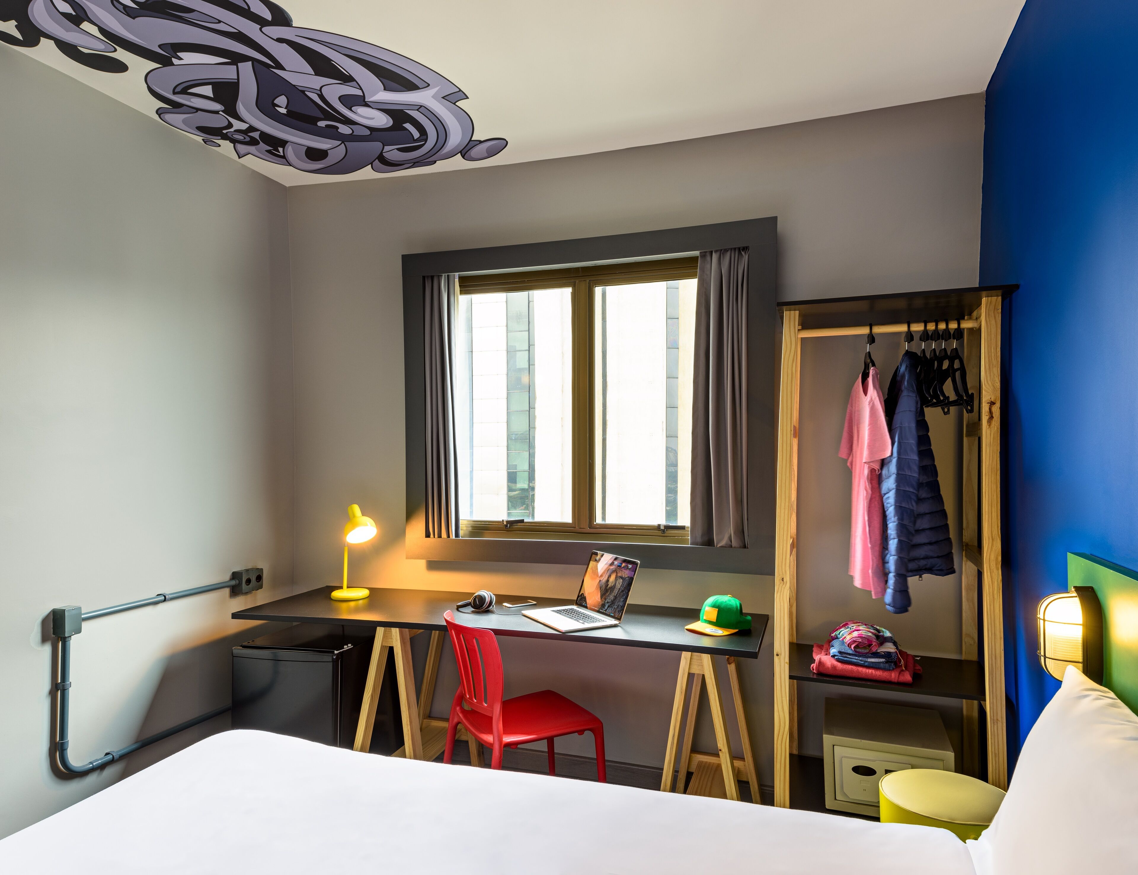 A compact hotel room with a desk, chair, and bed. A window with curtains, a coat rack with clothes, and a small bedside table with a lamp are visible.