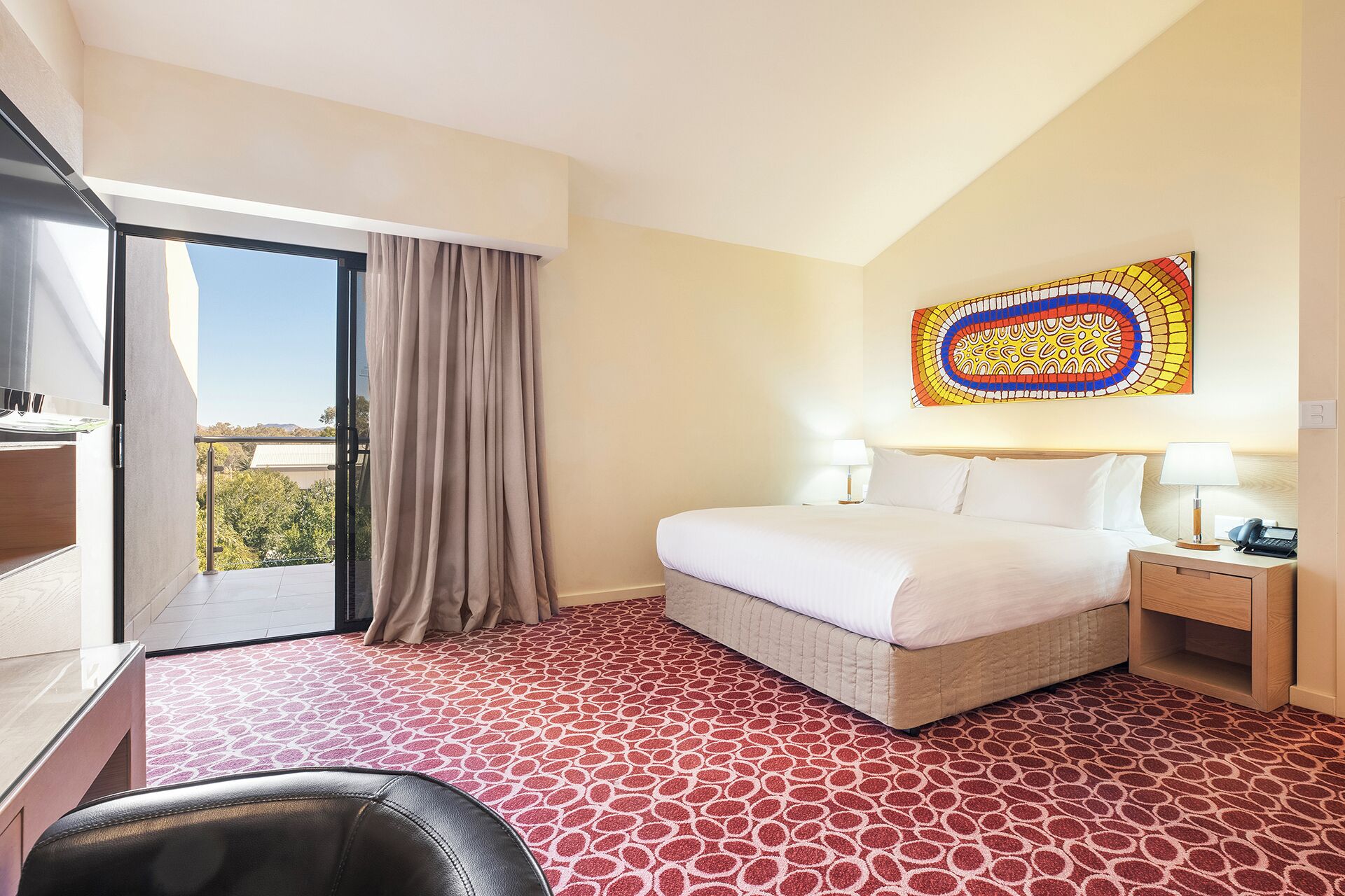 Une chambre d’hôtel avec un lit, un bureau, une chaise, une fenêtre avec des rideaux et une œuvre d’art colorée accrochée au mur.
