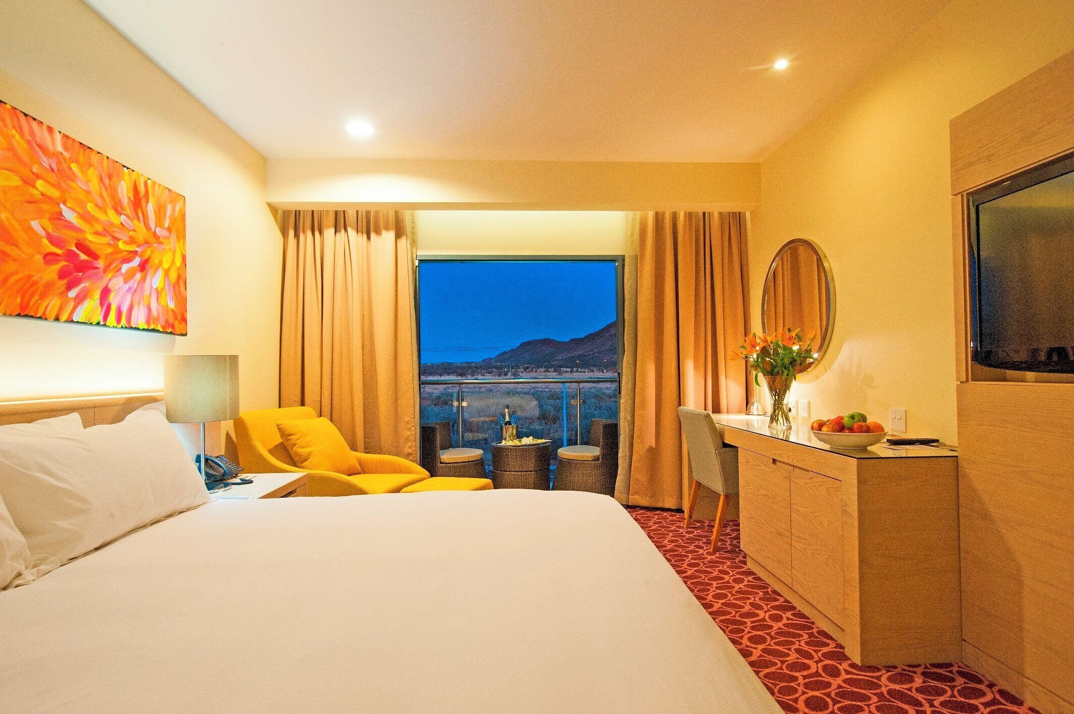 A hotel room with a large bed, a desk with a mirror, a chair, and a view of the mountains.