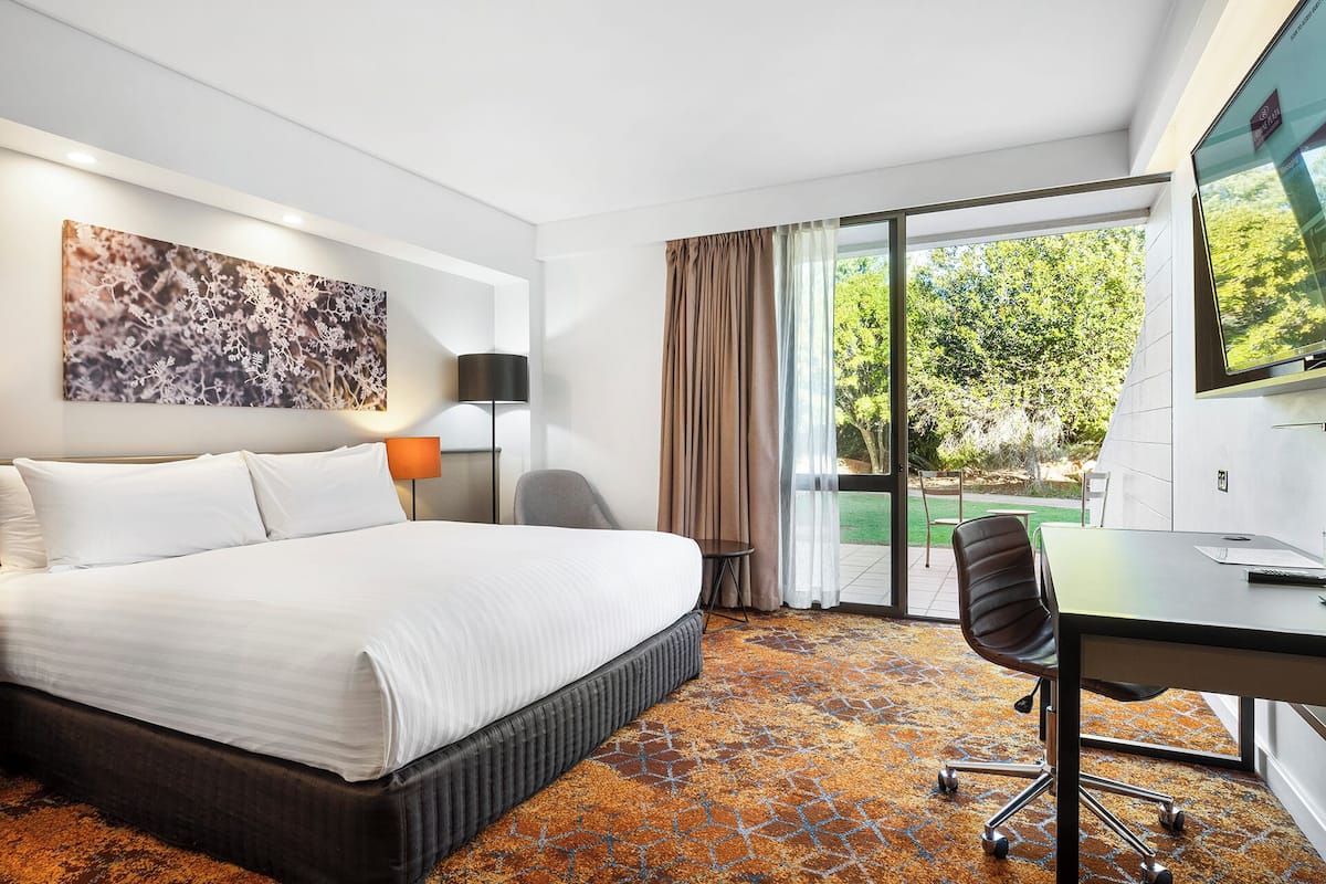 A hotel room with a bed, a desk, a chair, a lamp, and a large window with a view of greenery.