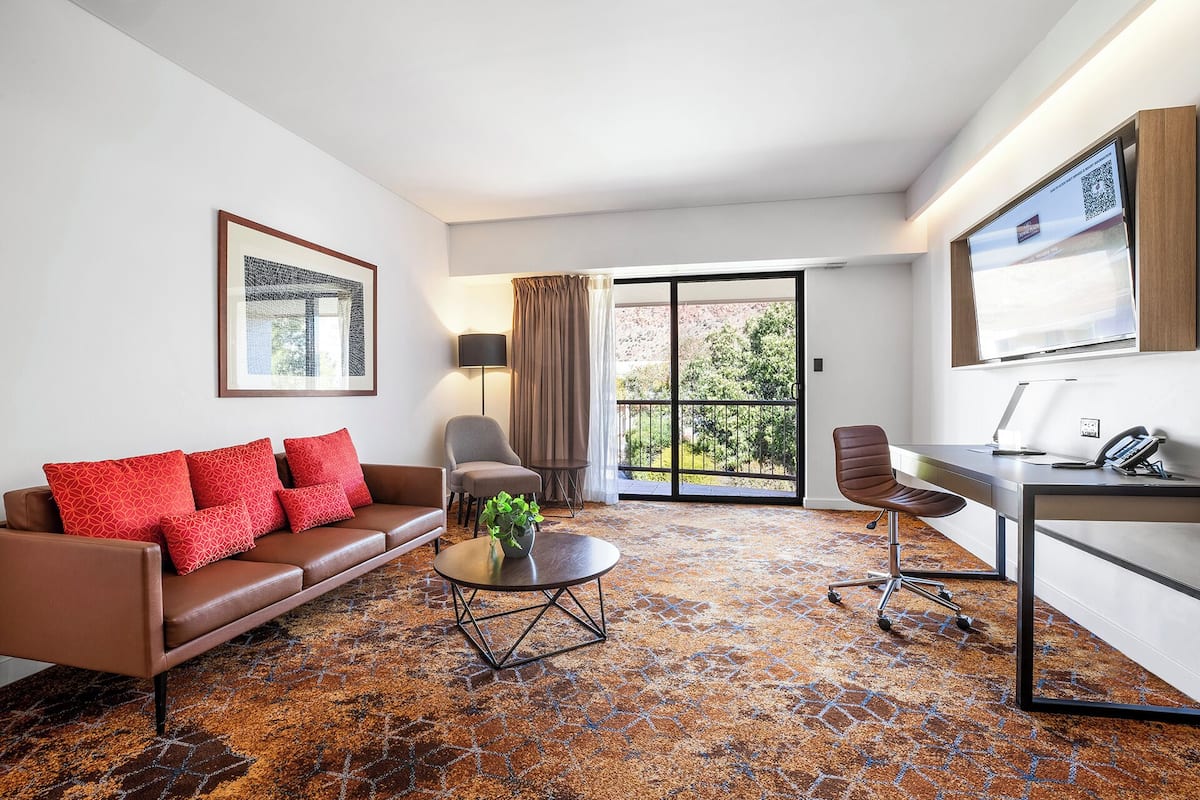A modern living room with a brown leather sofa, a round coffee table with a plant, a grey armchair, and a desk with a computer and phone. A large window offers a view of a garden and a mountain.