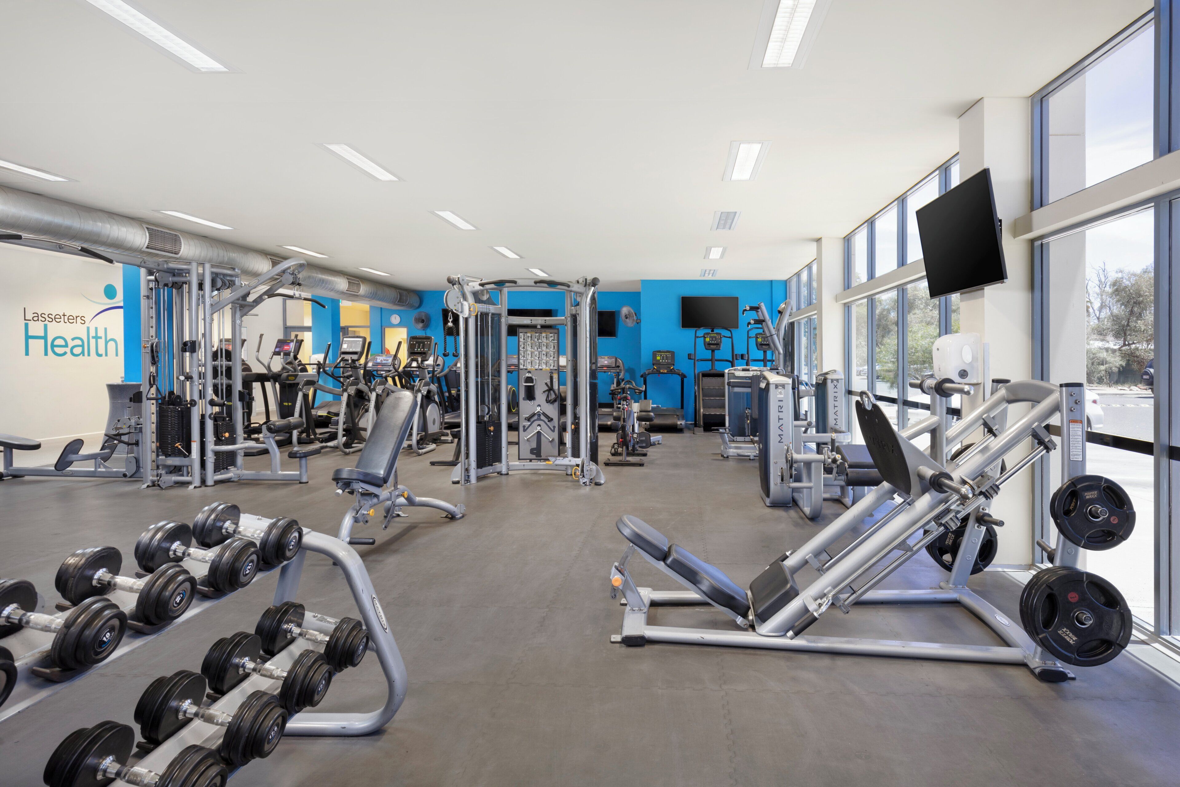 Une salle de sport équipée de divers appareils d’exercice, d’un téléviseur fixé au mur et de grandes fenêtres.