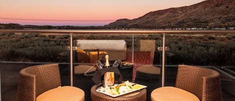 A balcony with wicker chairs and a table, overlooking a desert landscape with mountains in the distance.