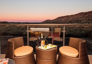 A balcony with wicker chairs and a table, overlooking a desert landscape with mountains in the distance.
