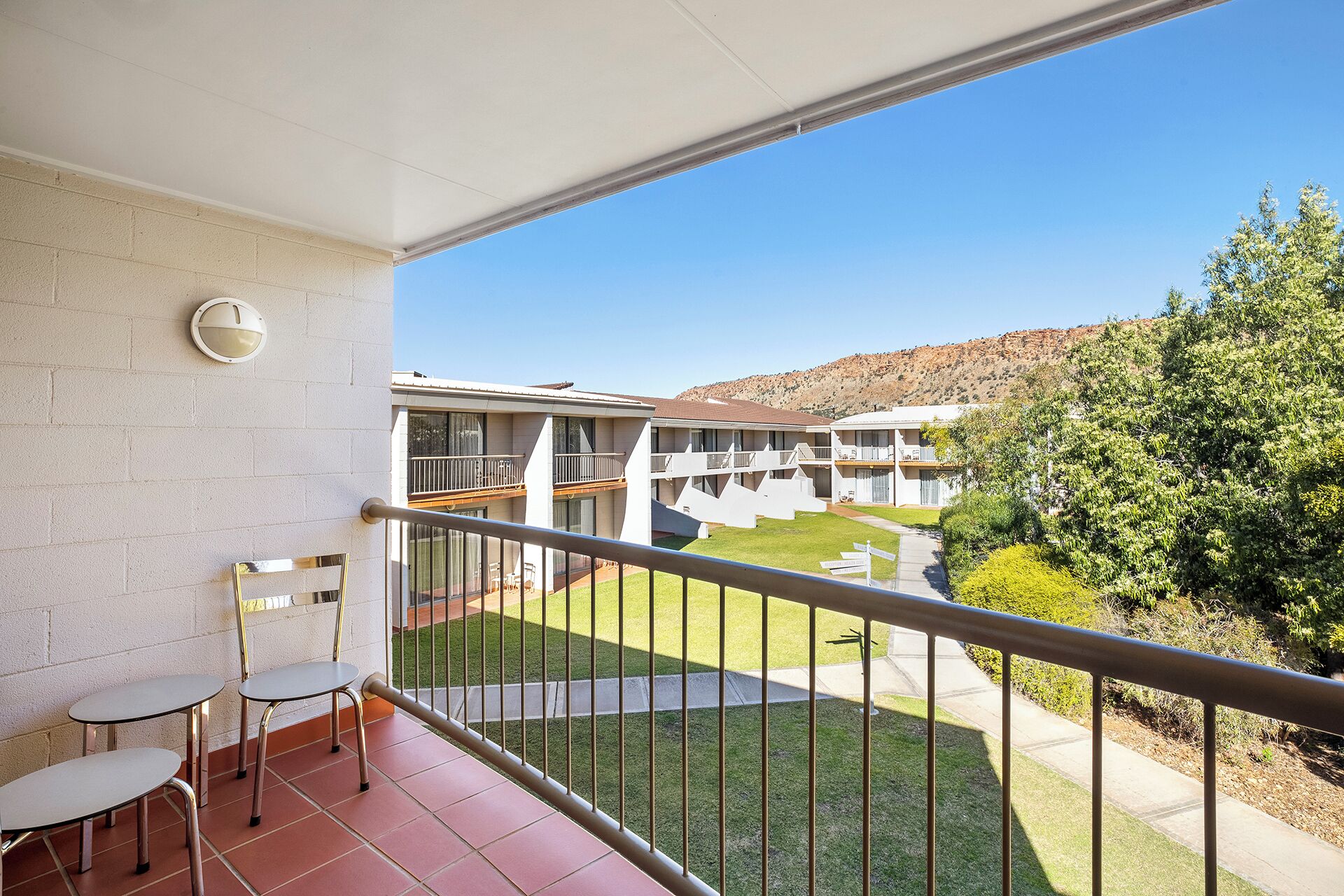 A balcony with two chairs, a small table, and a view of a building with multiple balconies and a grassy area.