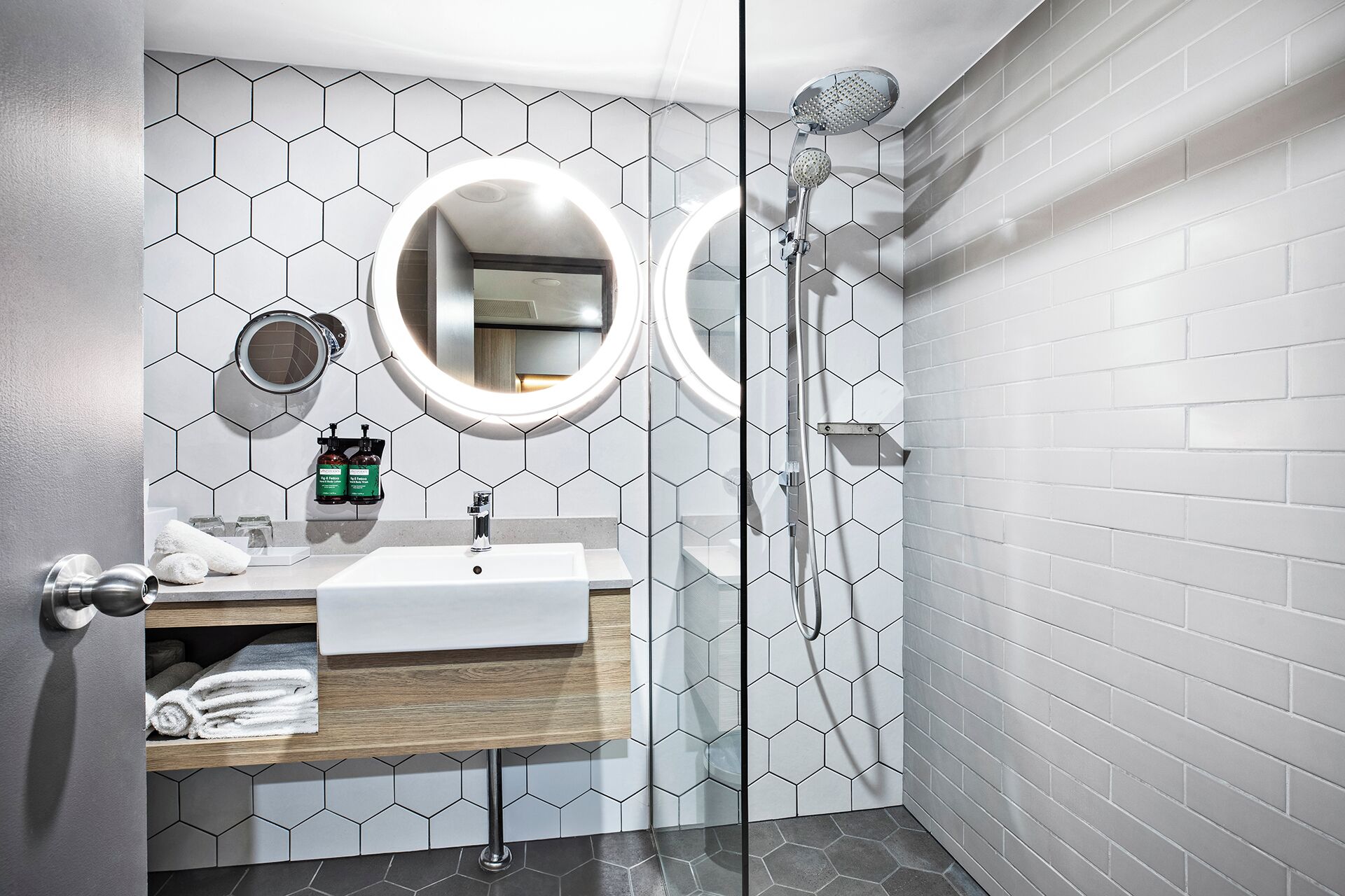 A modern bathroom with a white sink, wooden vanity, two round mirrors, and a shower with a handheld showerhead.