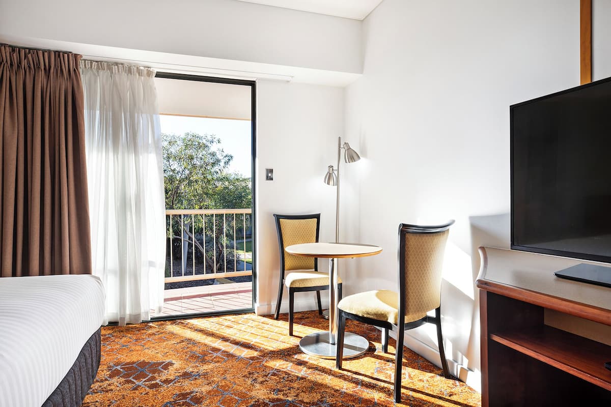 A hotel room with a bed, a small table with two chairs, a TV, and a balcony with a view of trees.