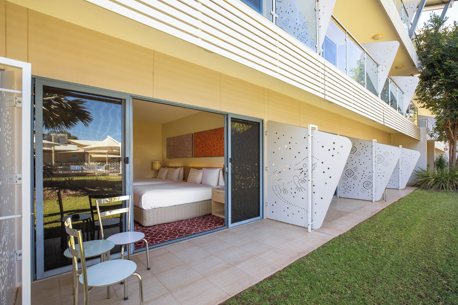 A hotel room with a bed, a desk, and a chair. There is a sliding glass door leading to a balcony with a table and chairs.