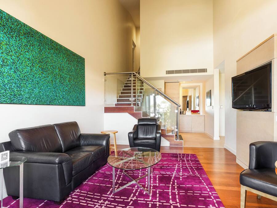 A modern living room with a black leather sofa, a glass coffee table, and a vibrant purple rug. A large green abstract painting adorns the wall, and a flat-screen TV is mounted on the wall.