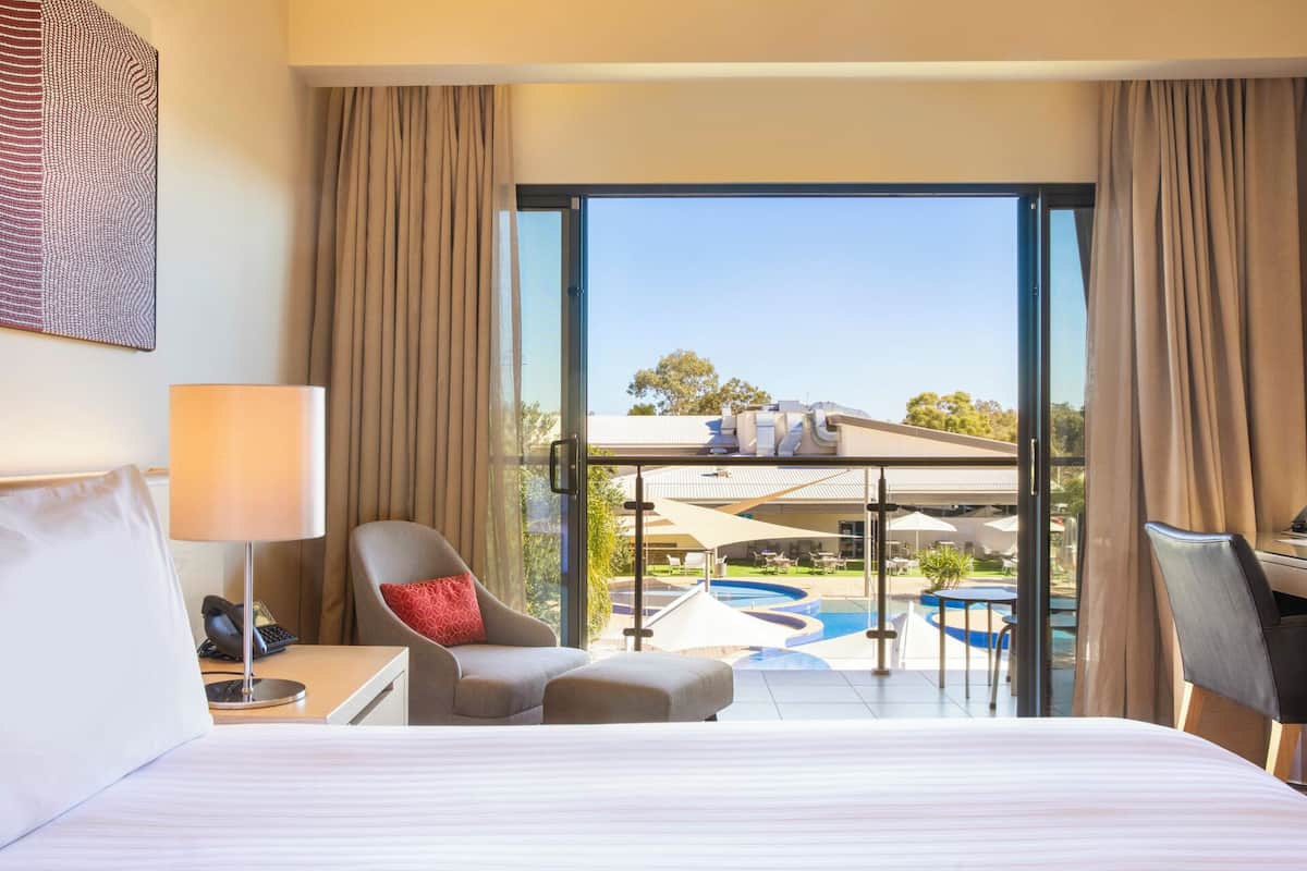 A hotel room with a bed, a desk, a chair, a lamp, and a view of an outdoor pool and lounge area.