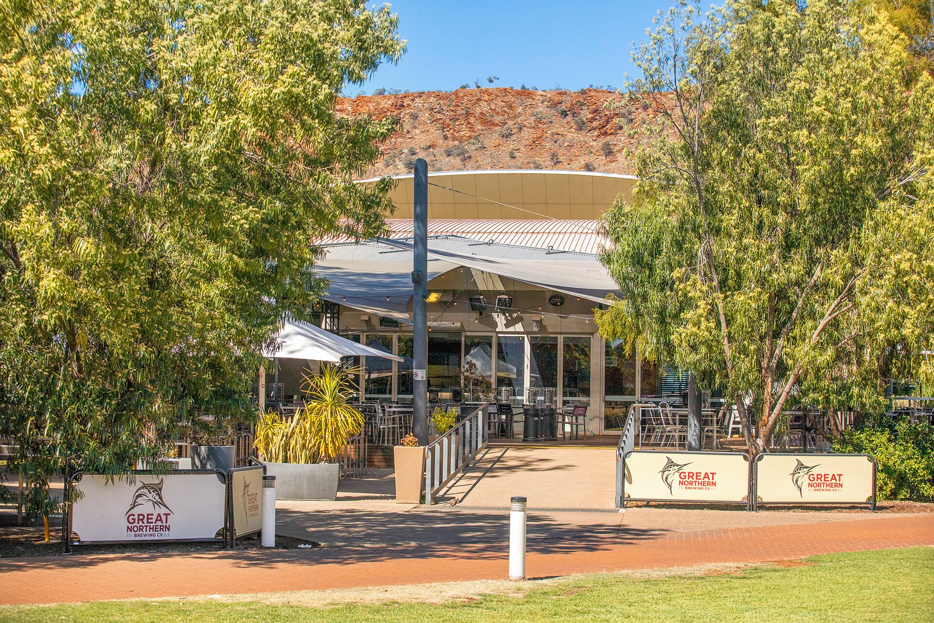 A building with a covered outdoor seating area, surrounded by trees and a hilly landscape in the background.