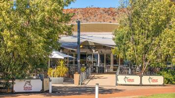 A building with a covered outdoor seating area, surrounded by trees and a hilly landscape in the background.