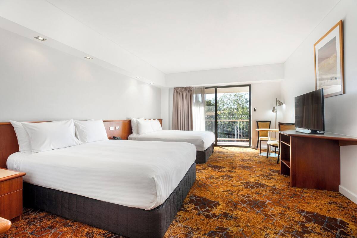 A hotel room with two beds, a desk, a TV, and a balcony with a view of trees.