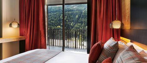 Une chambre avec une grande fenêtre, un lit recouvert d’une couette à carreaux, et une vue sur une forêt.