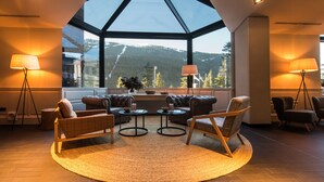 A modern interior with a high ceiling and large windows offering a mountain view. Furnishings include leather sofas, armchairs, and a round coffee table.