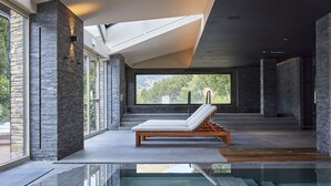 A modern indoor pool area with a lounge chair, stone walls, and large windows offering a view of greenery.