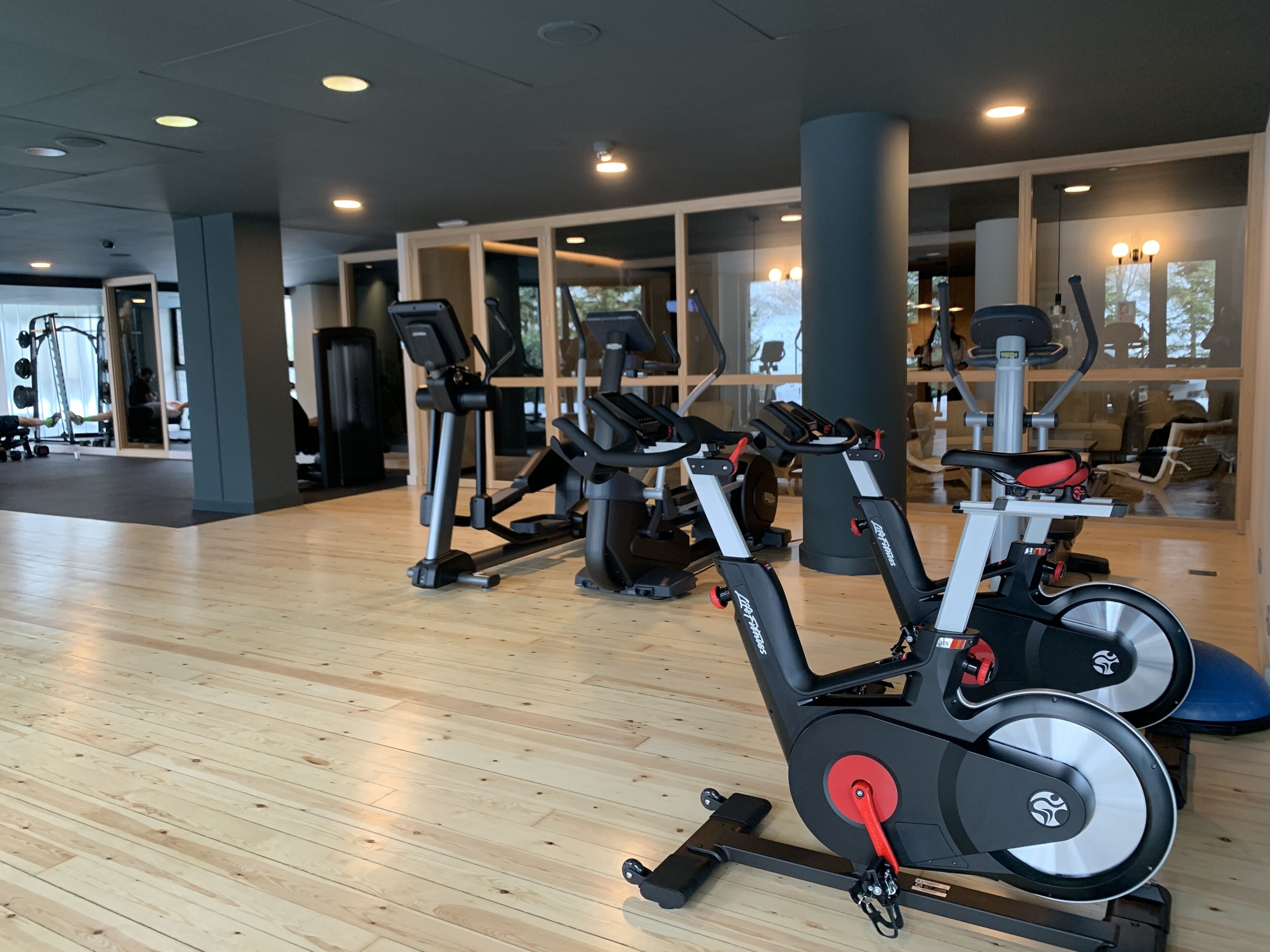 Un gimnasio con equipamiento de ejercicio, suelo de madera y ventanas amplias.