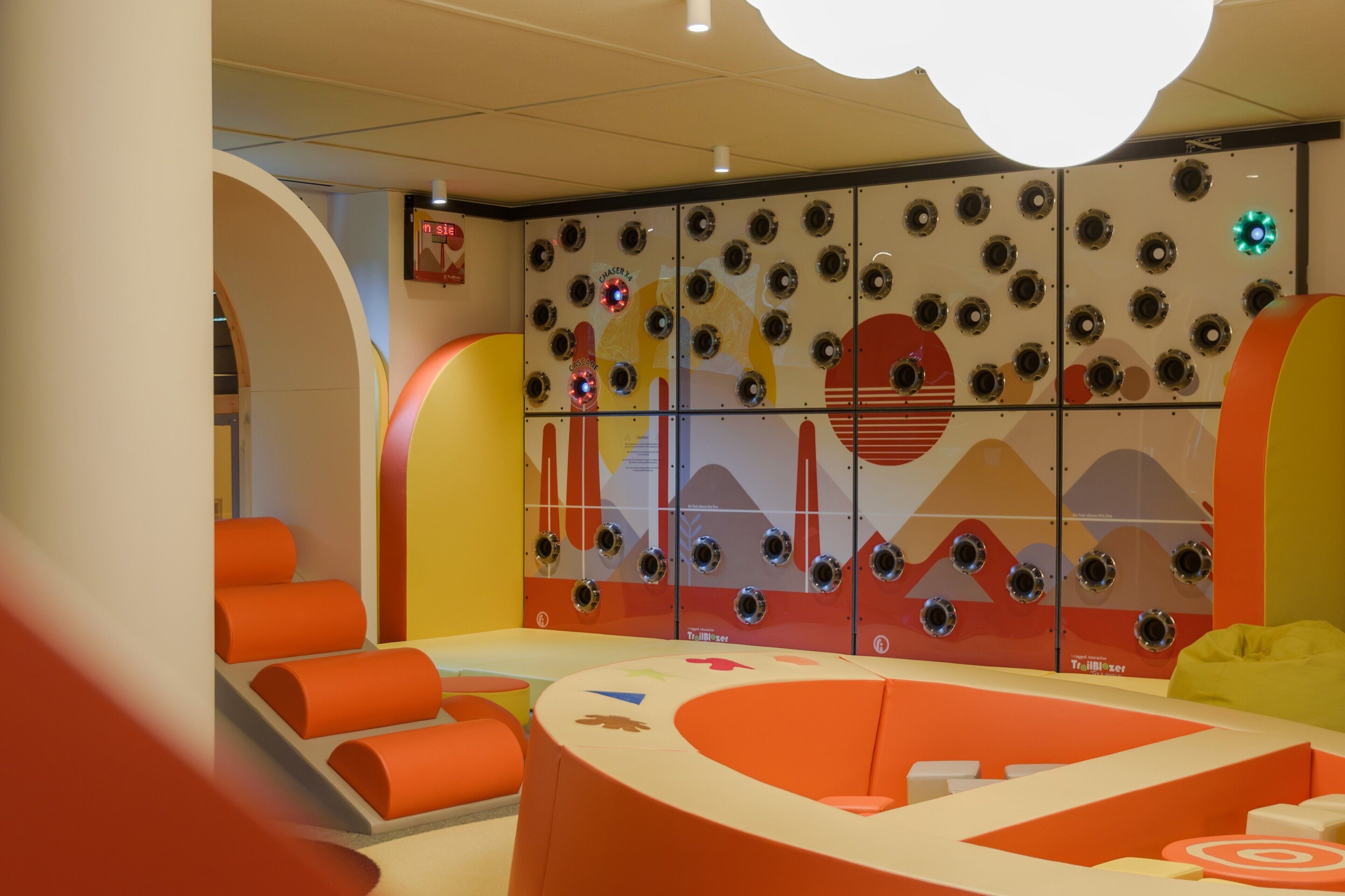 Zona infantil con muebles redondeados y coloridos, y una pared decorada con diseños circulares y un objetivo.