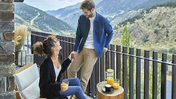 Una pareja en un balcón con una vista panorámica de las montañas, una mesa de madera con una taza de café y un vaso de zumo de naranja.