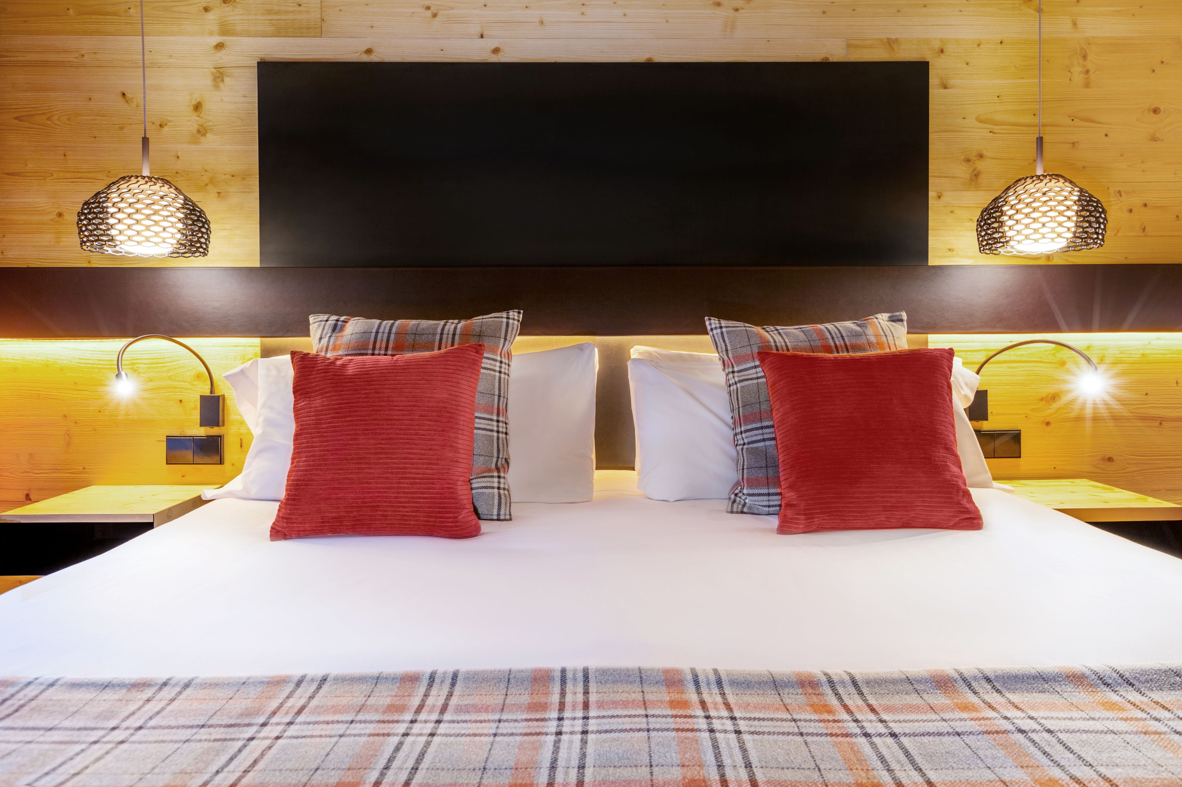 Una cama doble con dos almohadas rojas, un plaid y un televisor de pantalla plana grande montado en una pared de madera.