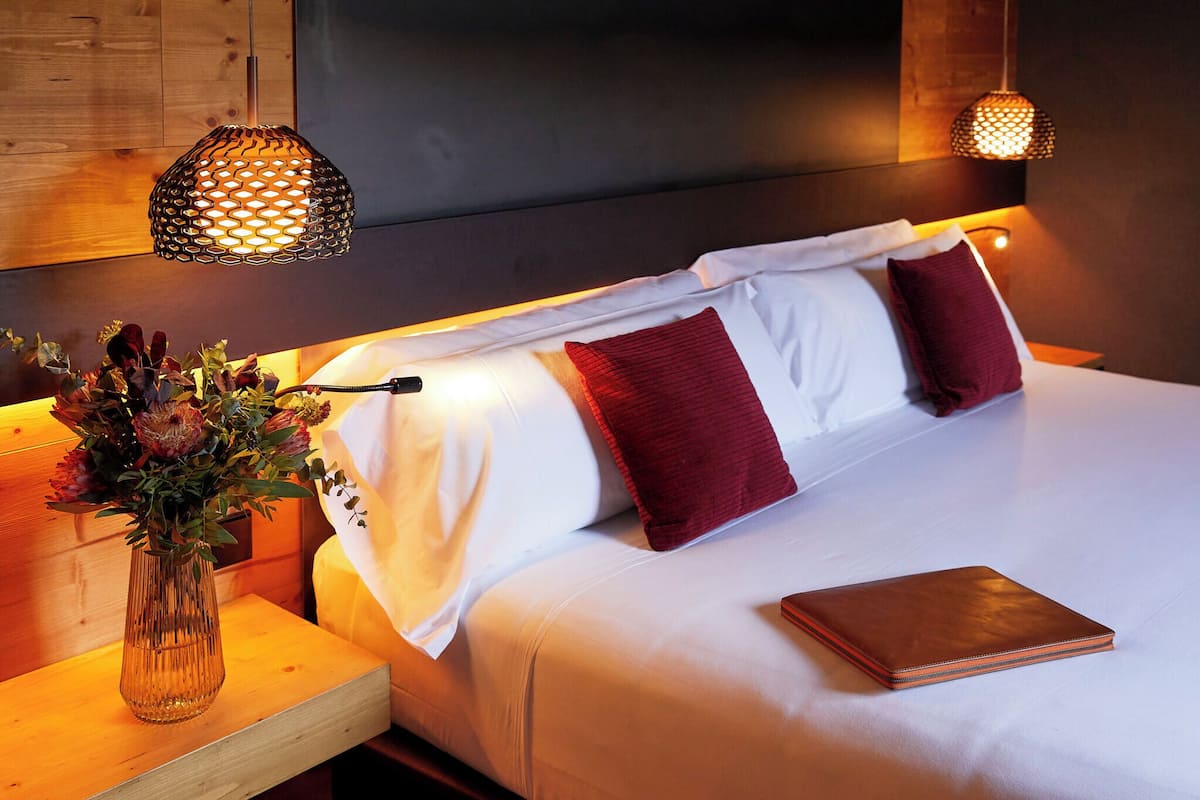 A neatly made bed with white and red pillows, a vase with flowers, and two hanging lamps.