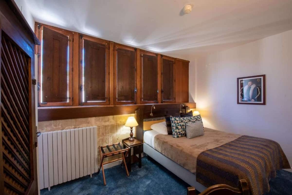 A bedroom with a bed, a nightstand, a lamp, a radiator, and wooden cabinets above the bed.