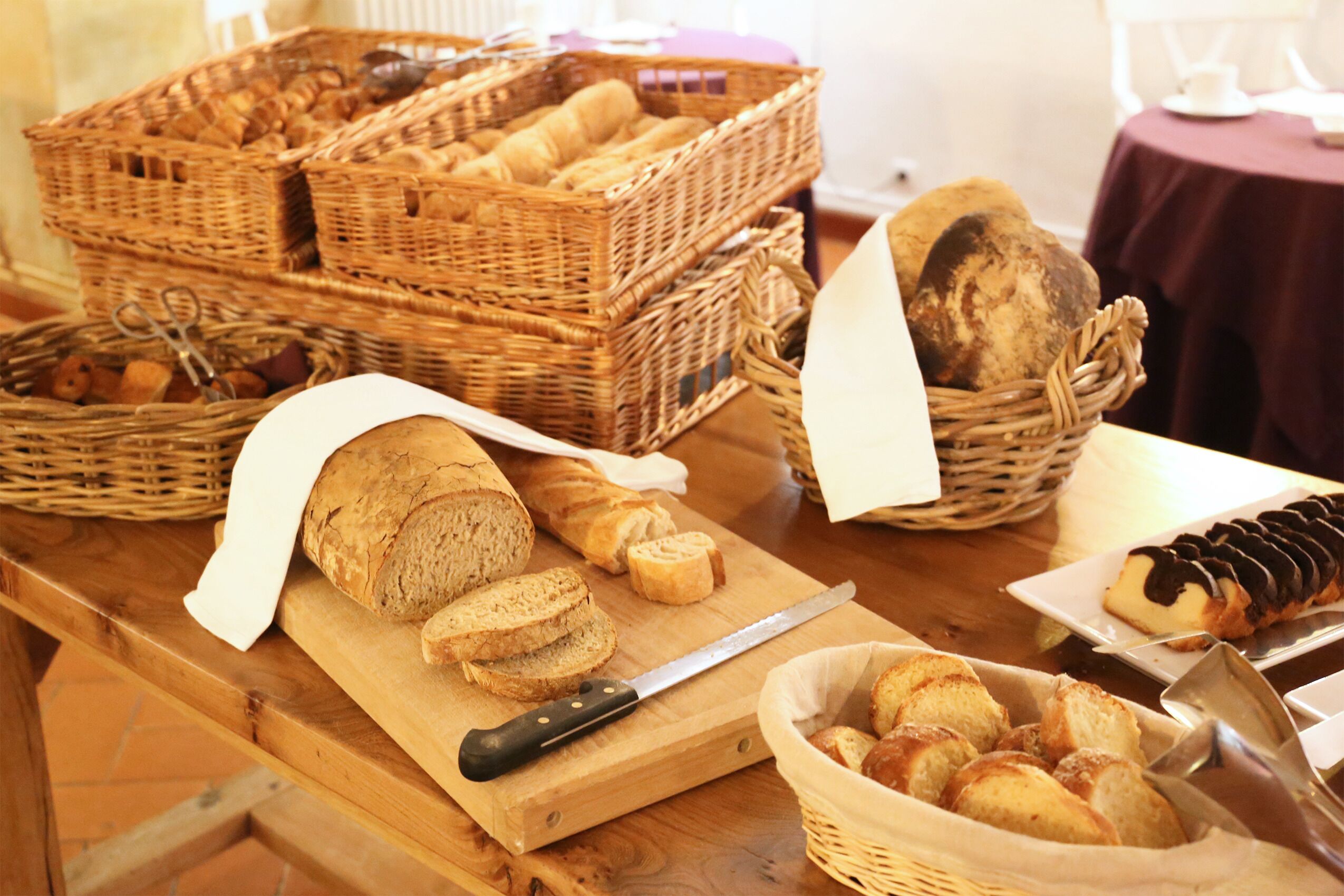 A wooden table with bread baskets, a loaf of sliced bread, a knife, and a plate with a chocolate-topped pastry.