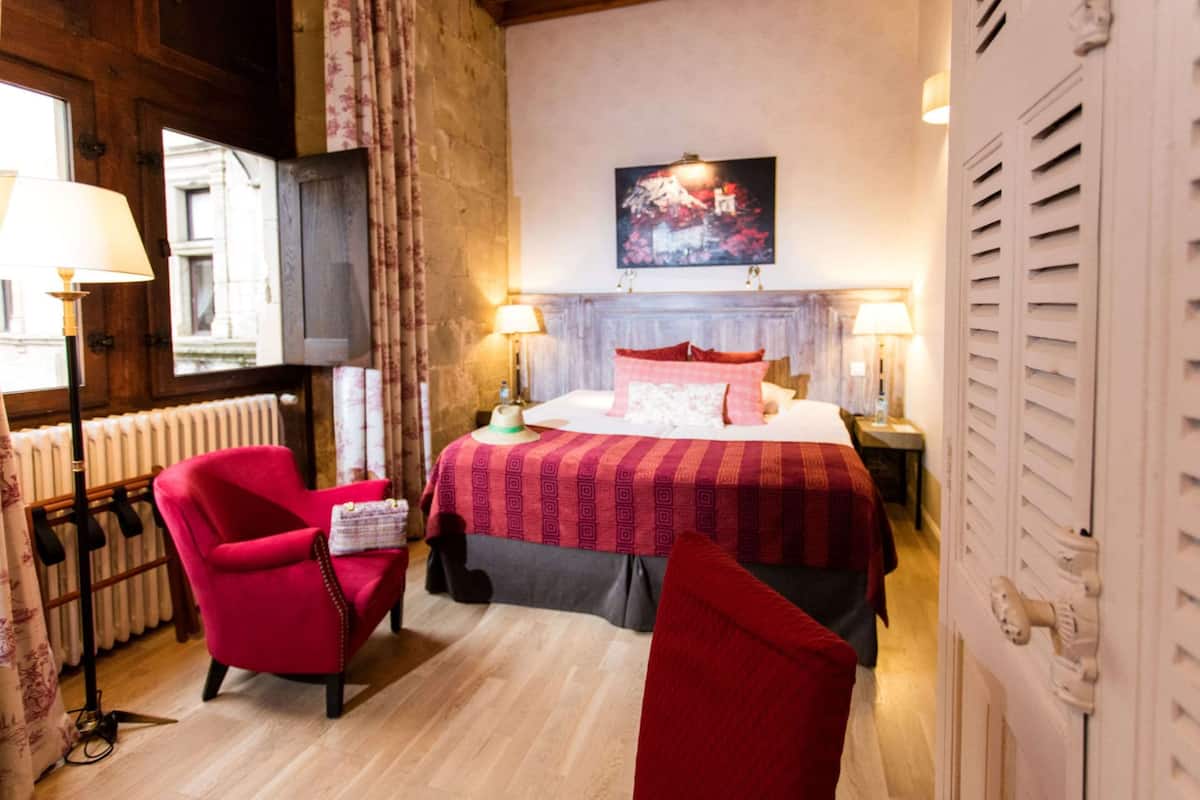A hotel room with a wooden floor, a red armchair, a bed with a patterned bedspread, and a painting on the wall.