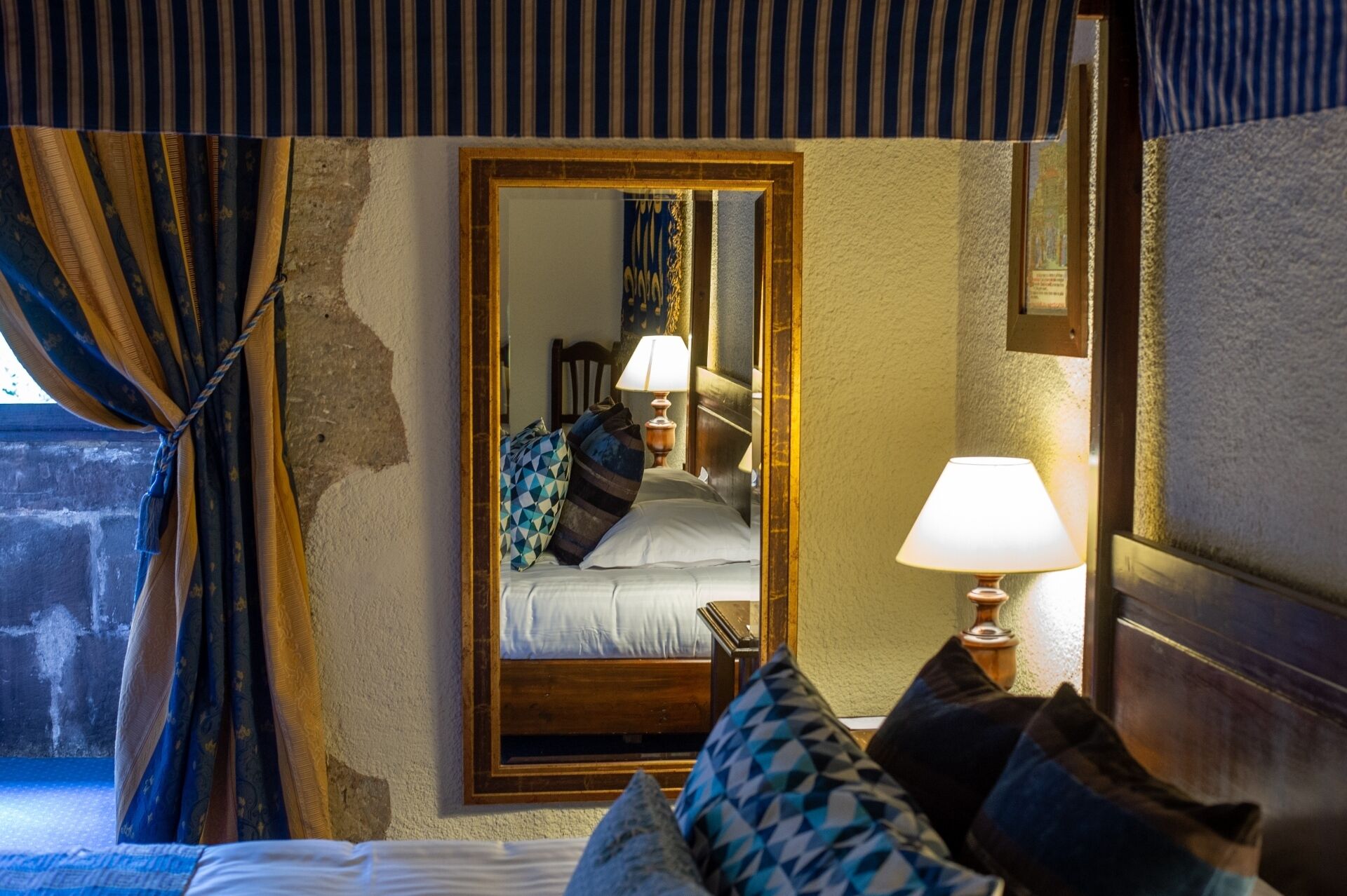 Habitación de hotel con cama, mesita de noche con lámpara, espejo y ventana con cortinas.
