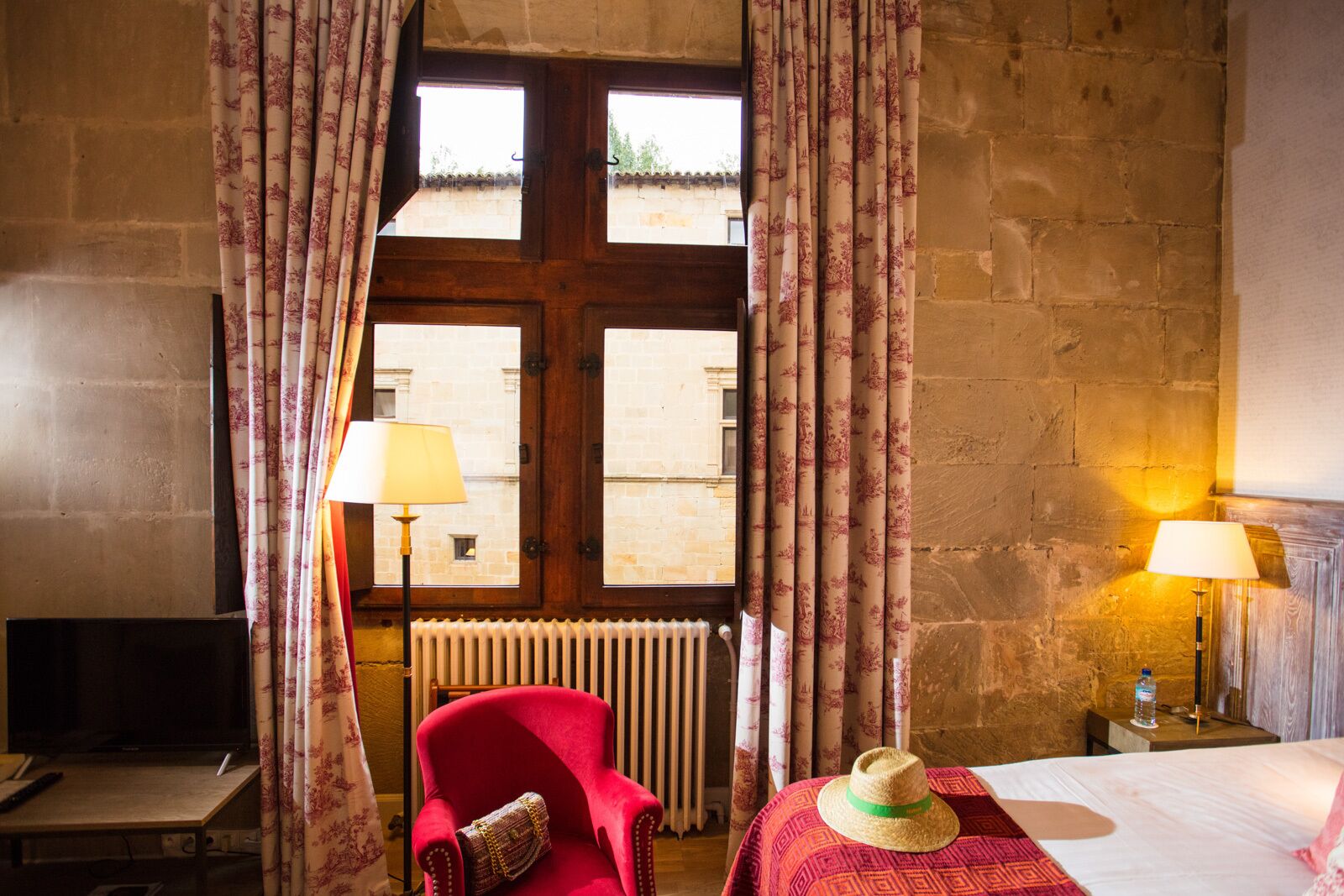 Una habitación de hotel con un sillón rojo, una cama con un sombrero de paja y dos lámparas.
