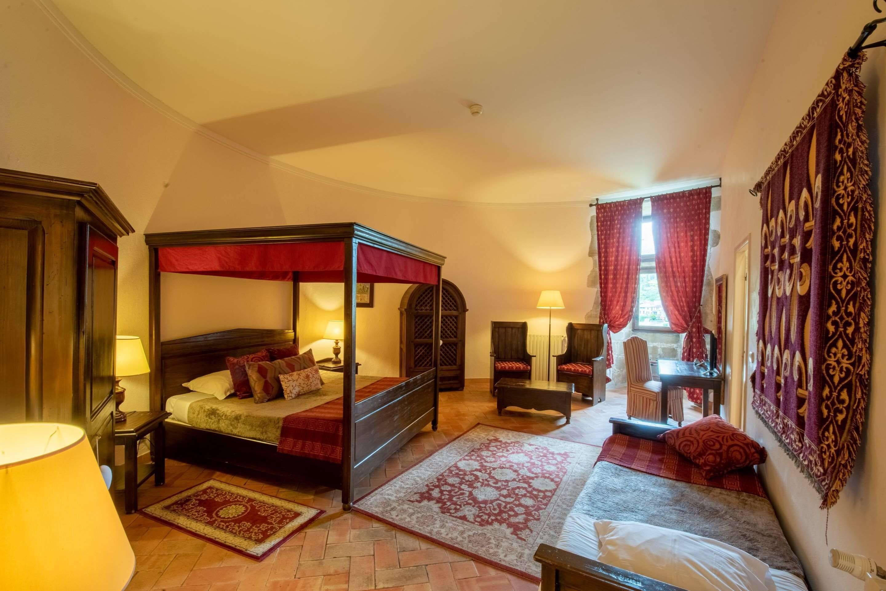 A four-poster bed with a red canopy, a wooden nightstand with a lamp, a patterned rug, and a small table with a chair.