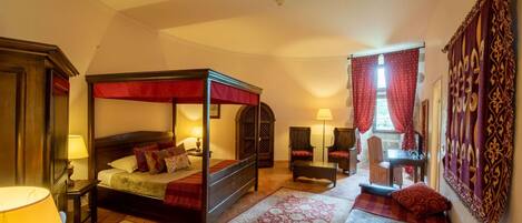 A four-poster bed with a red canopy, a wooden nightstand with a lamp, a patterned rug, and a small table with a chair.