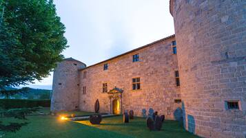 A stone building with a central entrance, a courtyard with seating, and a large tree to the left.