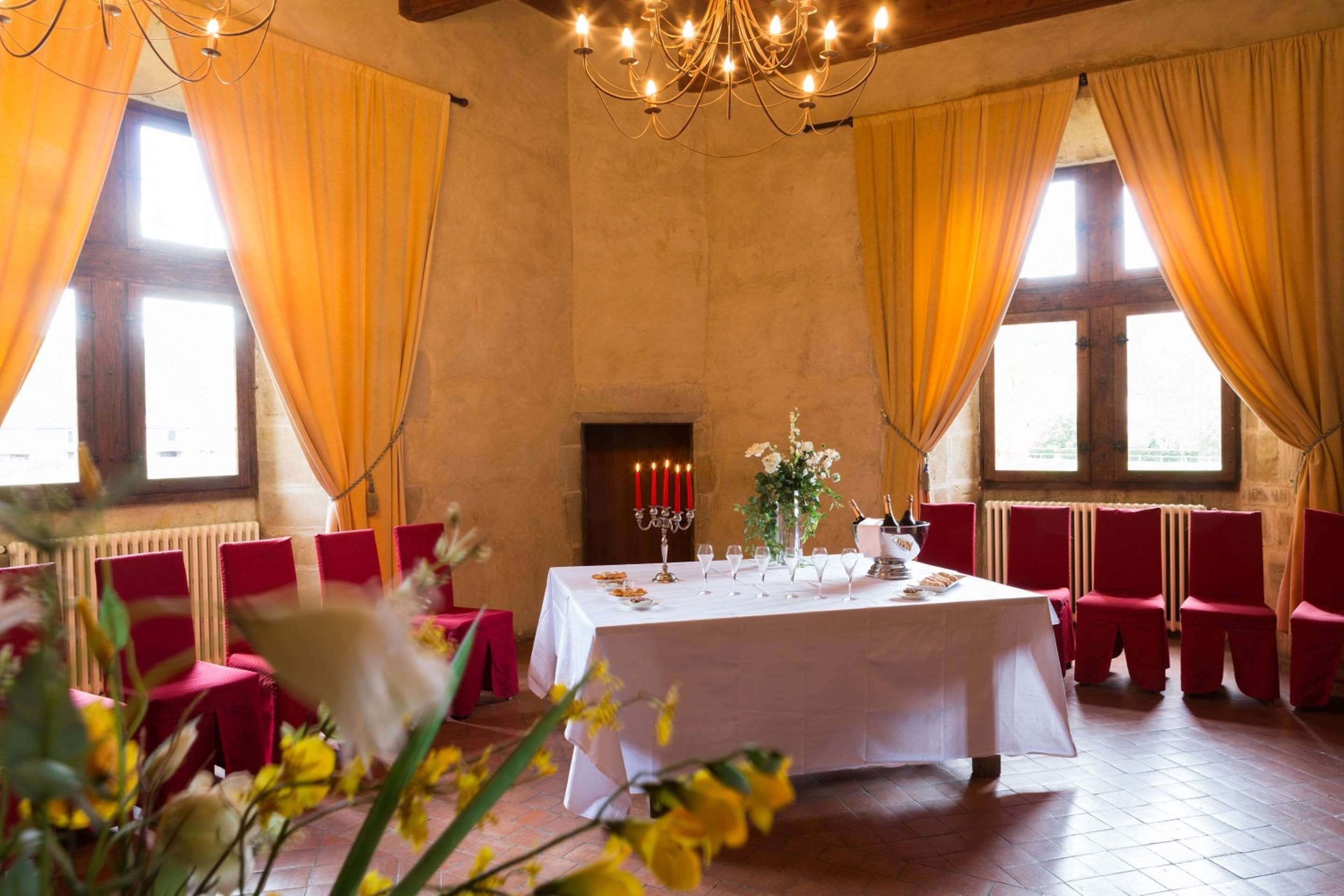 Uma sala de banquetes com uma longa mesa posta para refeição, cadeiras vermelhas, um lustre e grandes janelas com cortinas amarelas.